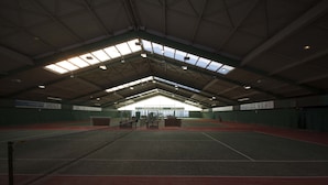 Una cancha de tenis interior con techo alto y tragaluces, rodeada de paredes verdes y piso rojo.