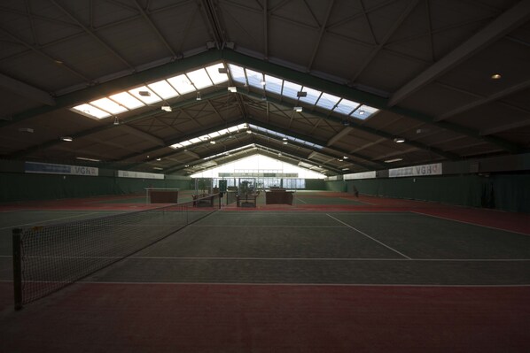 An indoor tennis court with a high ceiling and skylights, surrounded by green walls and a red floor.