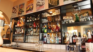 Zona de bar con estantes llenos de botellas y copas, un espejo y varios carteles en la pared.