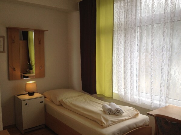 A hotel room with a bed, a nightstand with a lamp, a mirror, a window with sheer curtains, and a wooden wardrobe.