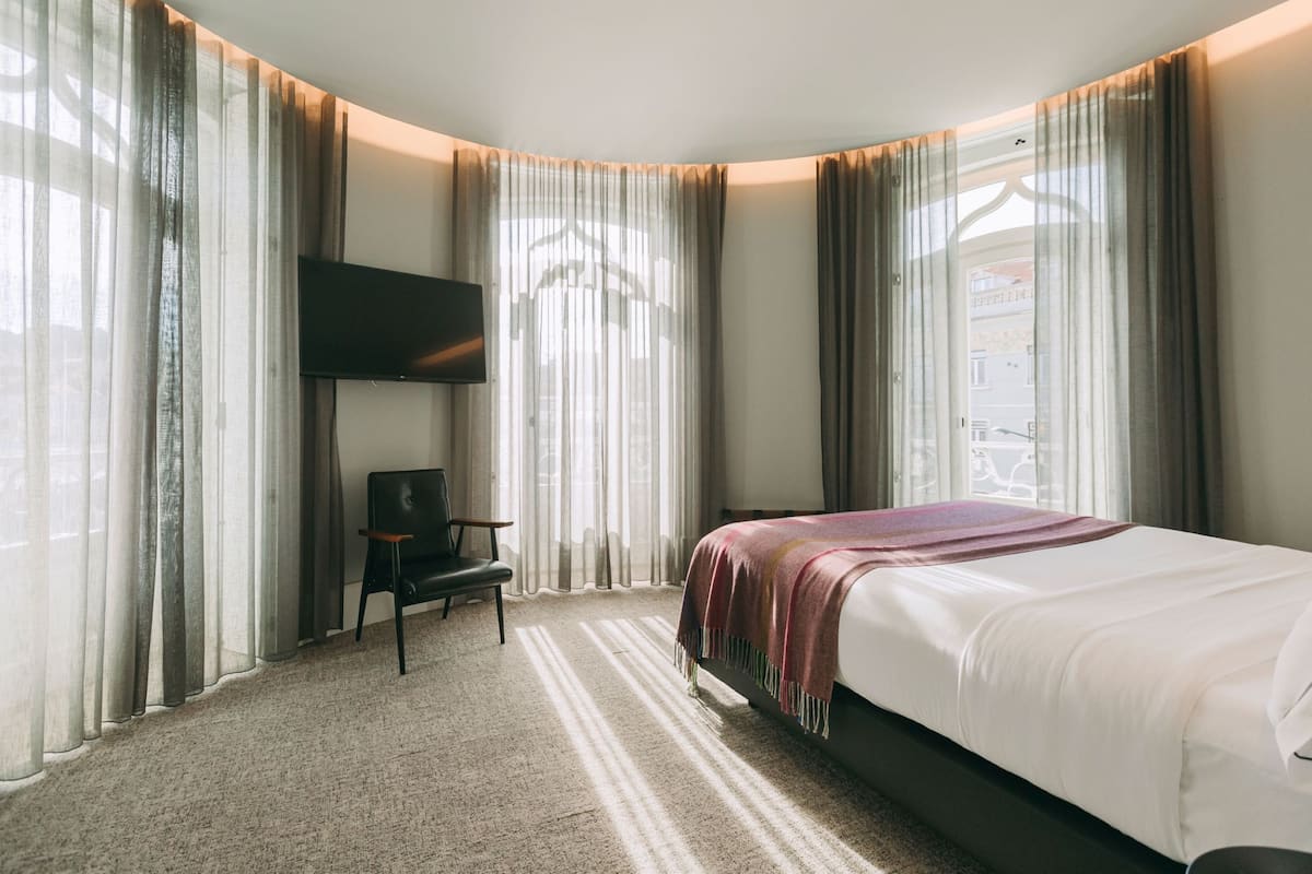 A hotel room with a large bed, a flat-screen TV mounted on the wall, a chair, and sheer curtains.