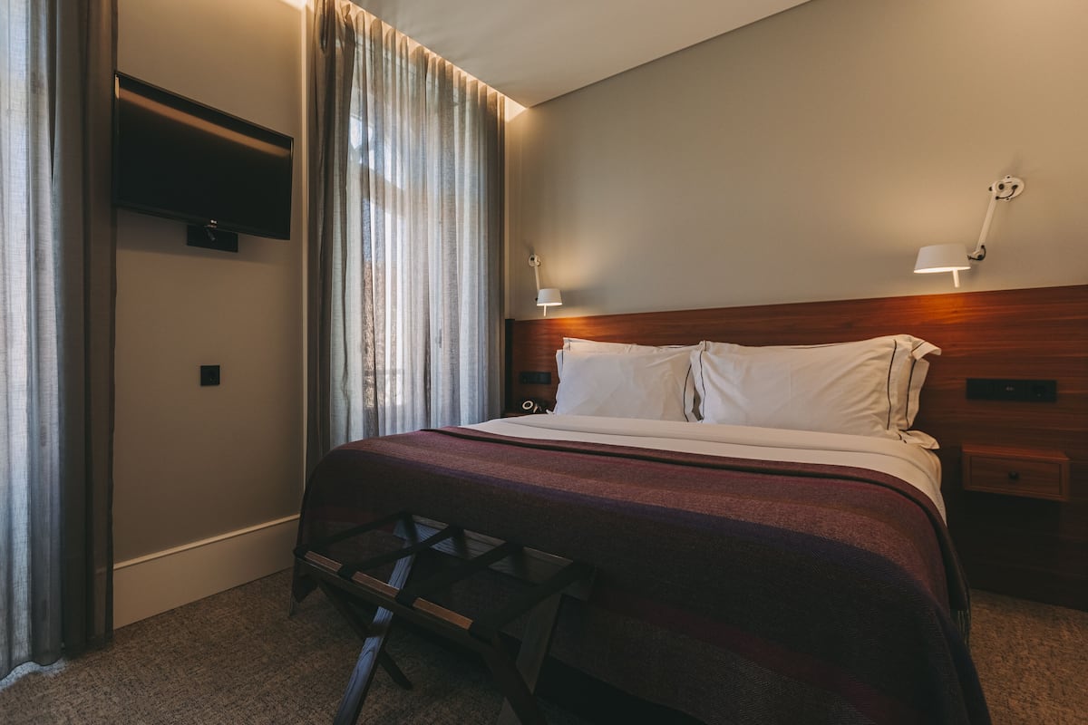 A hotel room with a bed, a TV mounted on the wall, a nightstand with a lamp, and a window with curtains.