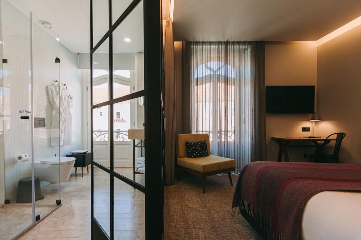 A hotel room with a glass shower enclosure, a bed with a red blanket, a chair, a desk, and a TV on the wall.