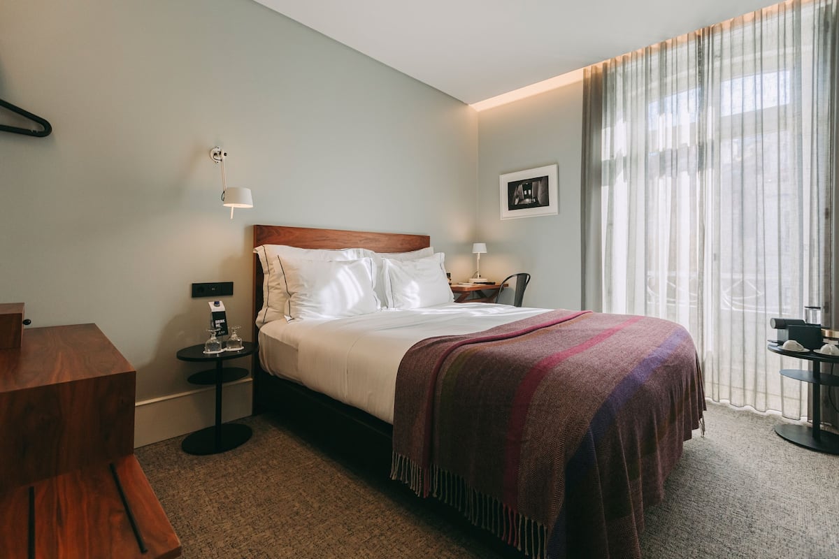 A hotel room with a bed, a nightstand, a wooden desk, a chair, a small table with a lamp, and a window with sheer curtains.
