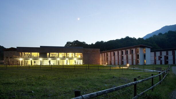 Un edificio moderno con múltiples ventanas y una entrada luminosa, rodeado de césped y un sendero.