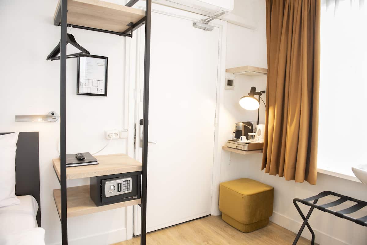 A compact hotel room with a bed, a desk with a lamp, a wardrobe, a small stool, and a hanging coat rack.