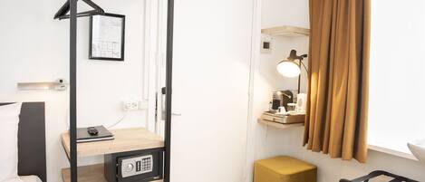 A compact hotel room with a bed, a desk with a lamp, a wardrobe, a small stool, and a hanging coat rack.