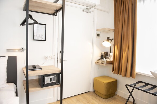 A compact hotel room with a bed, a desk with a lamp, a wardrobe, a small stool, and a hanging coat rack.