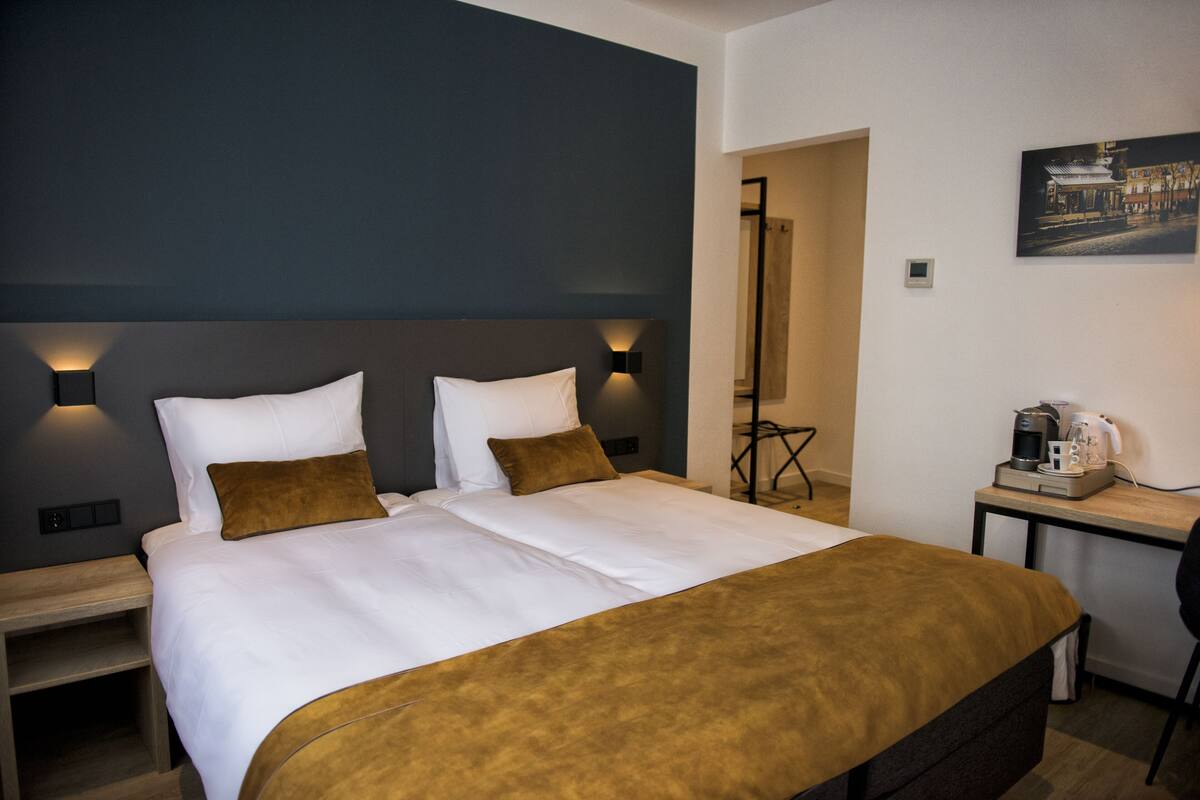 A double bed with white linens and two brown pillows, a bedside table with a lamp, and a small table with a coffee maker and a cup.