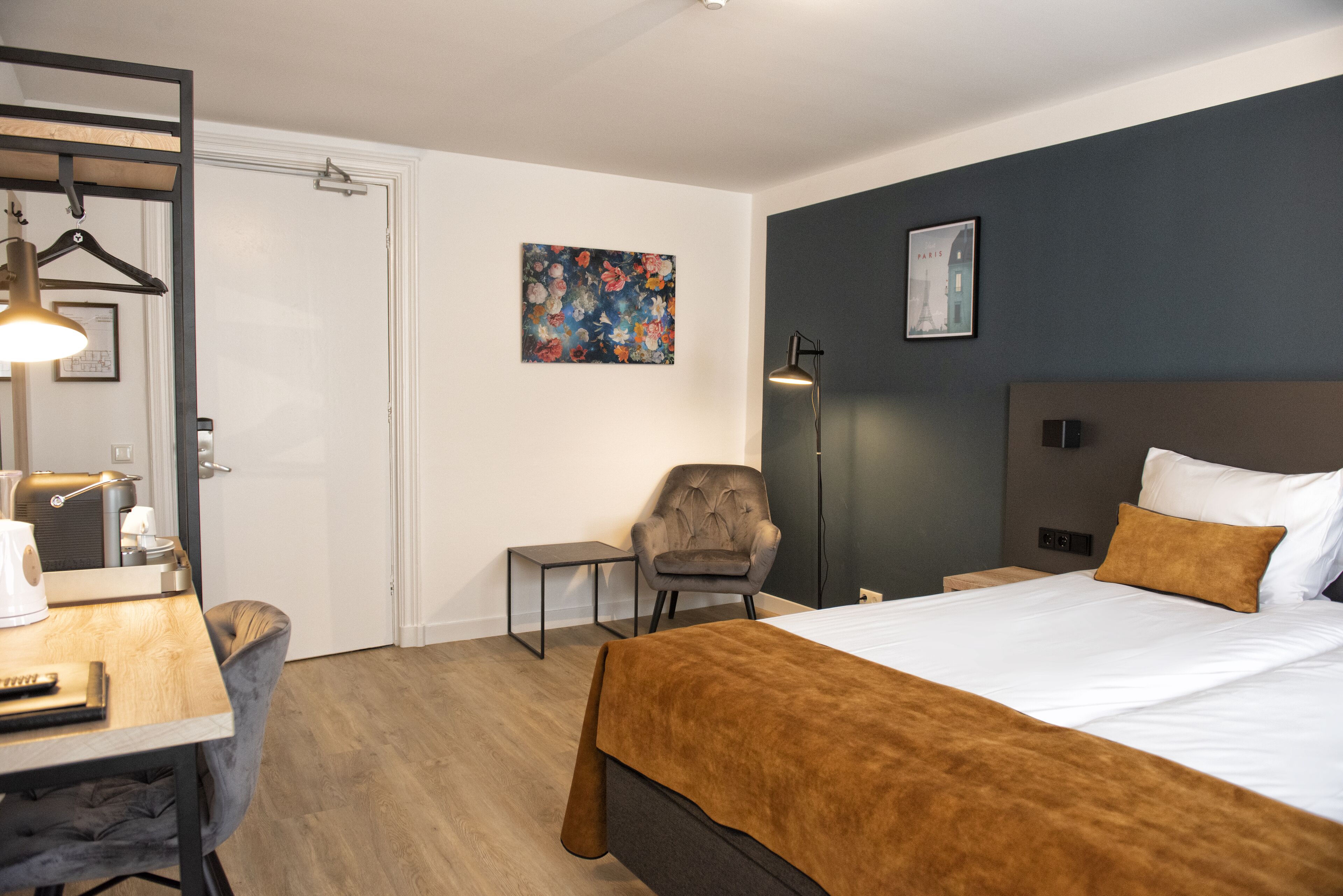 A hotel room with a bed, a desk, a chair, a bedside table, a lamp, and artwork on the walls.