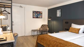 Ett hotellrum med en säng, ett skrivbord, en stol, ett sängbord, en lampa och konstverk på väggarna.