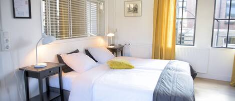 Habitación de hotel ordenada con cama, mesita de noche, lámpara y ventana con persianas.