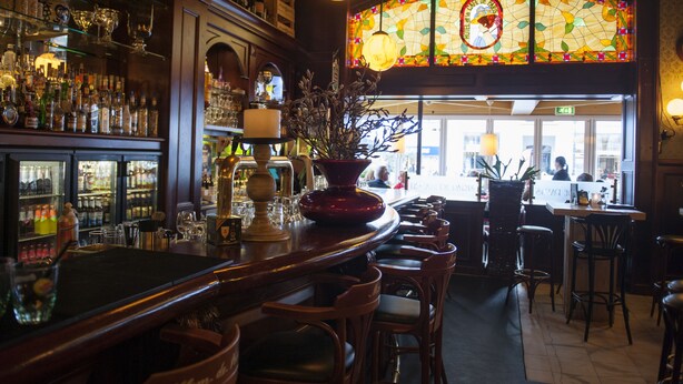 Zona de bar con un amplio ventanal de vitral, taburetes y una variedad de botellas y vasos en estanterías.