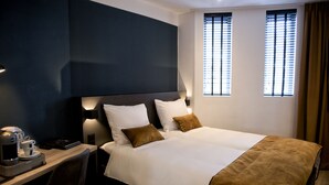 Habitación de hotel con una cama grande, dos mesitas de noche, un escritorio con cafetera y dos ventanas con persianas.