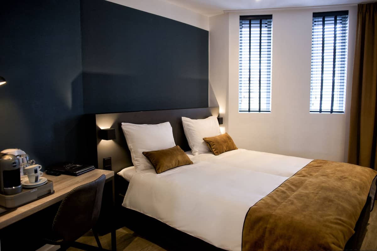 A hotel room with a large bed, two bedside tables, a desk with a coffee maker, and two windows with blinds.
