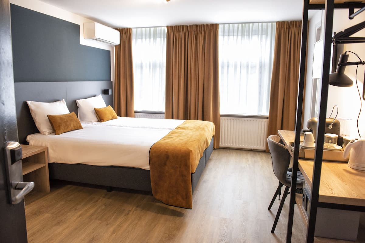 A modern hotel room with a large bed, a desk, and a chair. There is a window with curtains, a radiator, and a desk lamp.