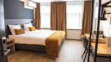 Habitación de hotel moderna con una cama grande, un escritorio y una silla. Se ve una ventana con cortinas, un radiador y una lámpara de escritorio.