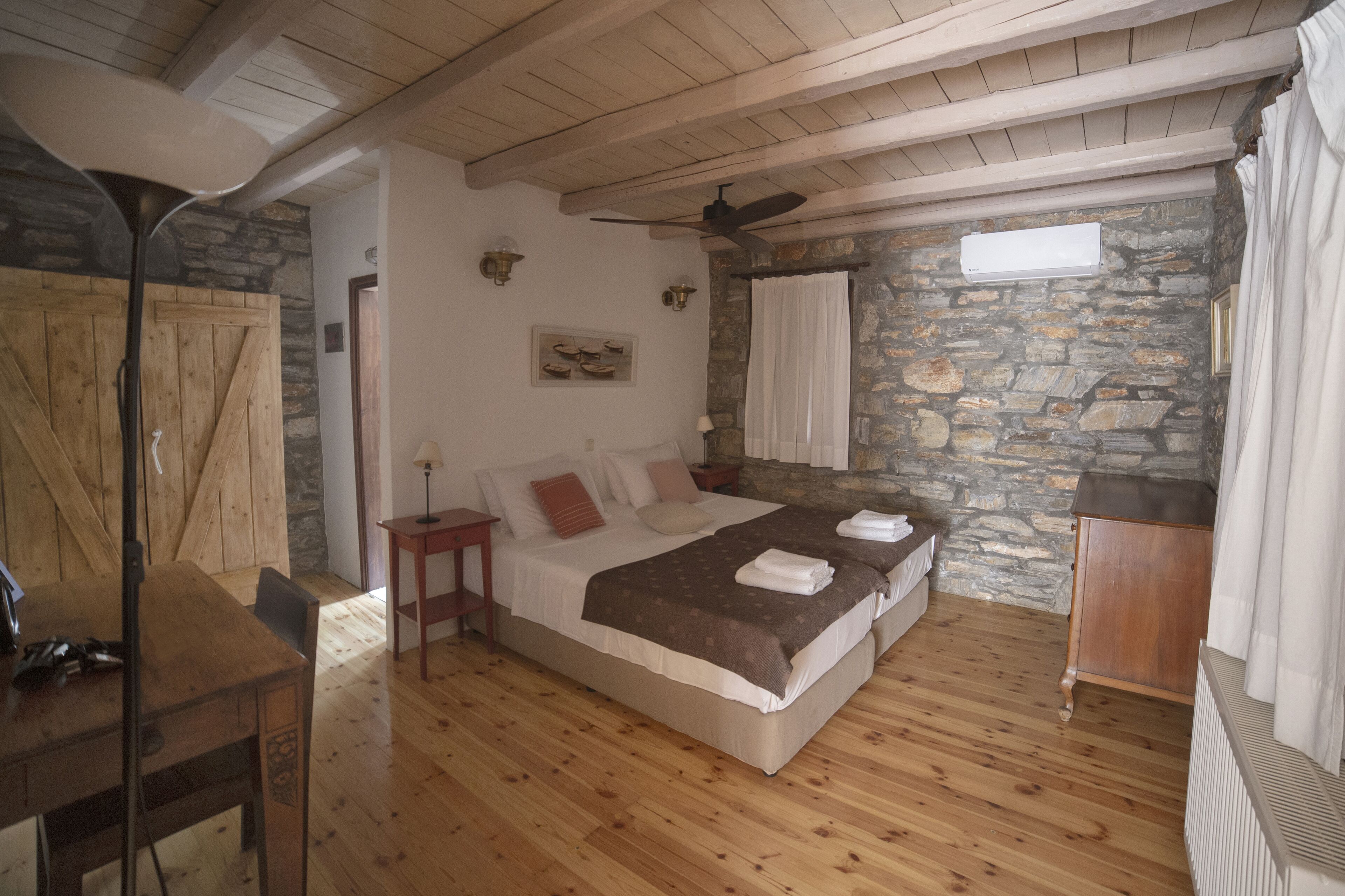 Un dormitorio con piso de madera, cama con manta marrón, ventilador de techo y pared de piedra.