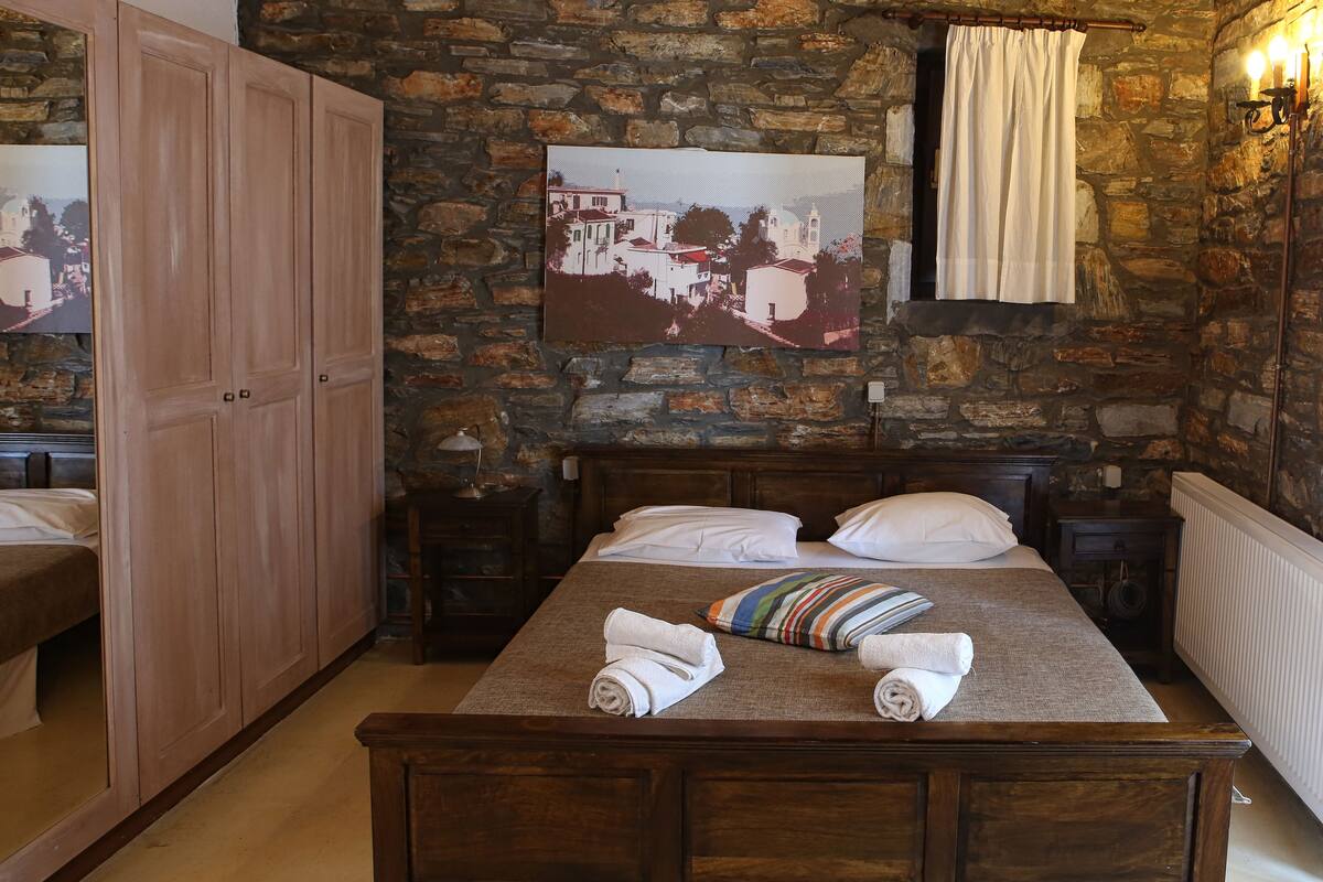 A bedroom with a stone wall, wooden bed frame, and a painting of a village scene.