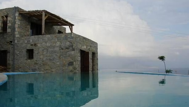 A stone building with a pool extending to the water's edge, a clear sky, and a palm tree in the distance. - Pyrgos Ikaria Traditional Village (Ikaria)