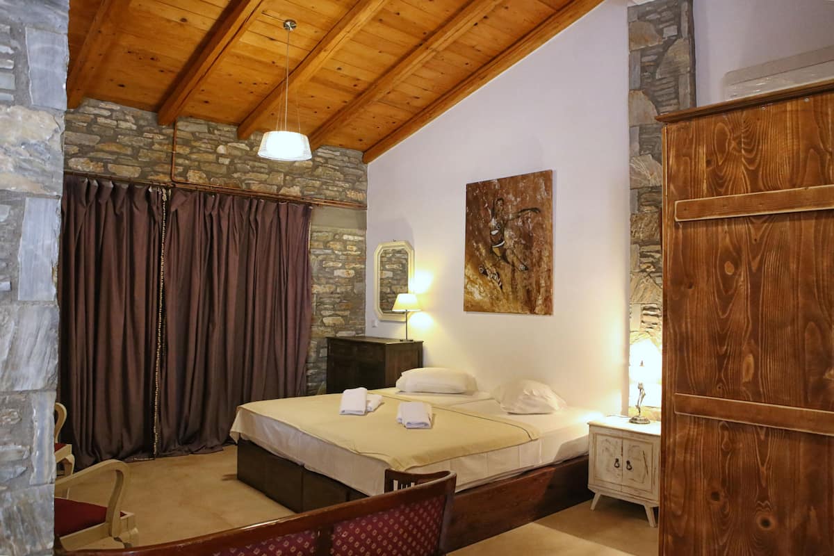 A bedroom with a stone wall, wooden ceiling beams, a bed with white linens, a nightstand with a lamp, and a wooden wardrobe.