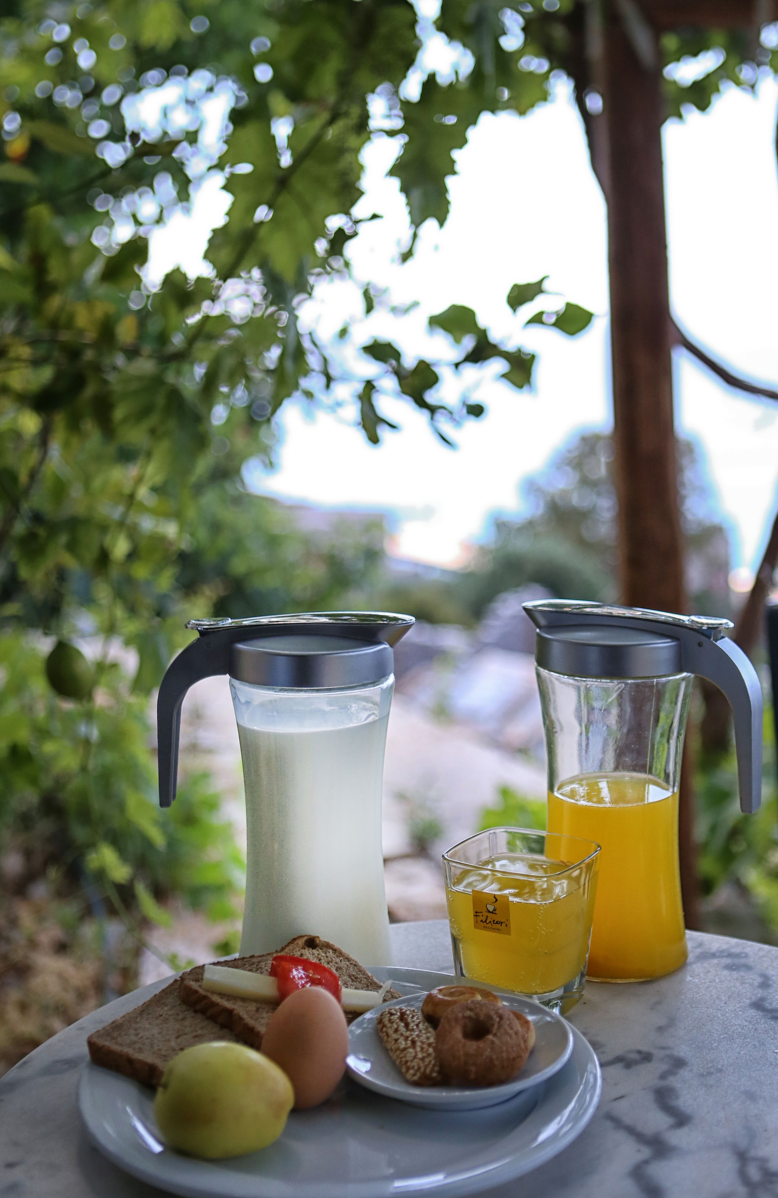 A table with a pitcher of milk, a glass of juice, toast with cheese and tomato, an egg, and a small plate with a lemon and a pastry.