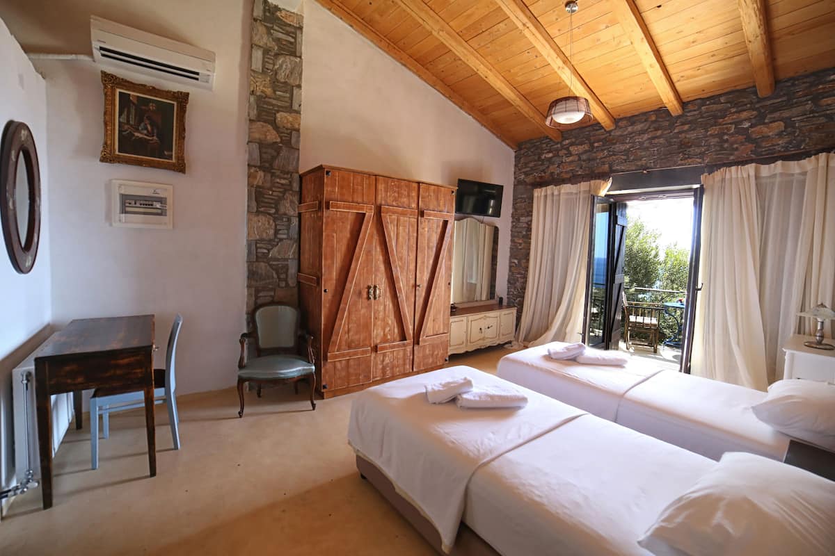 A room with two beds, a wooden wardrobe, a desk with a chair, a television, and a stone wall.