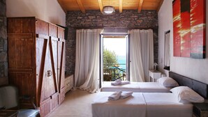 A bedroom with two beds, a wooden wardrobe, a stone wall, and a view of the sea through a large window. - Pyrgos Ikaria Traditional Village (Ikaria)