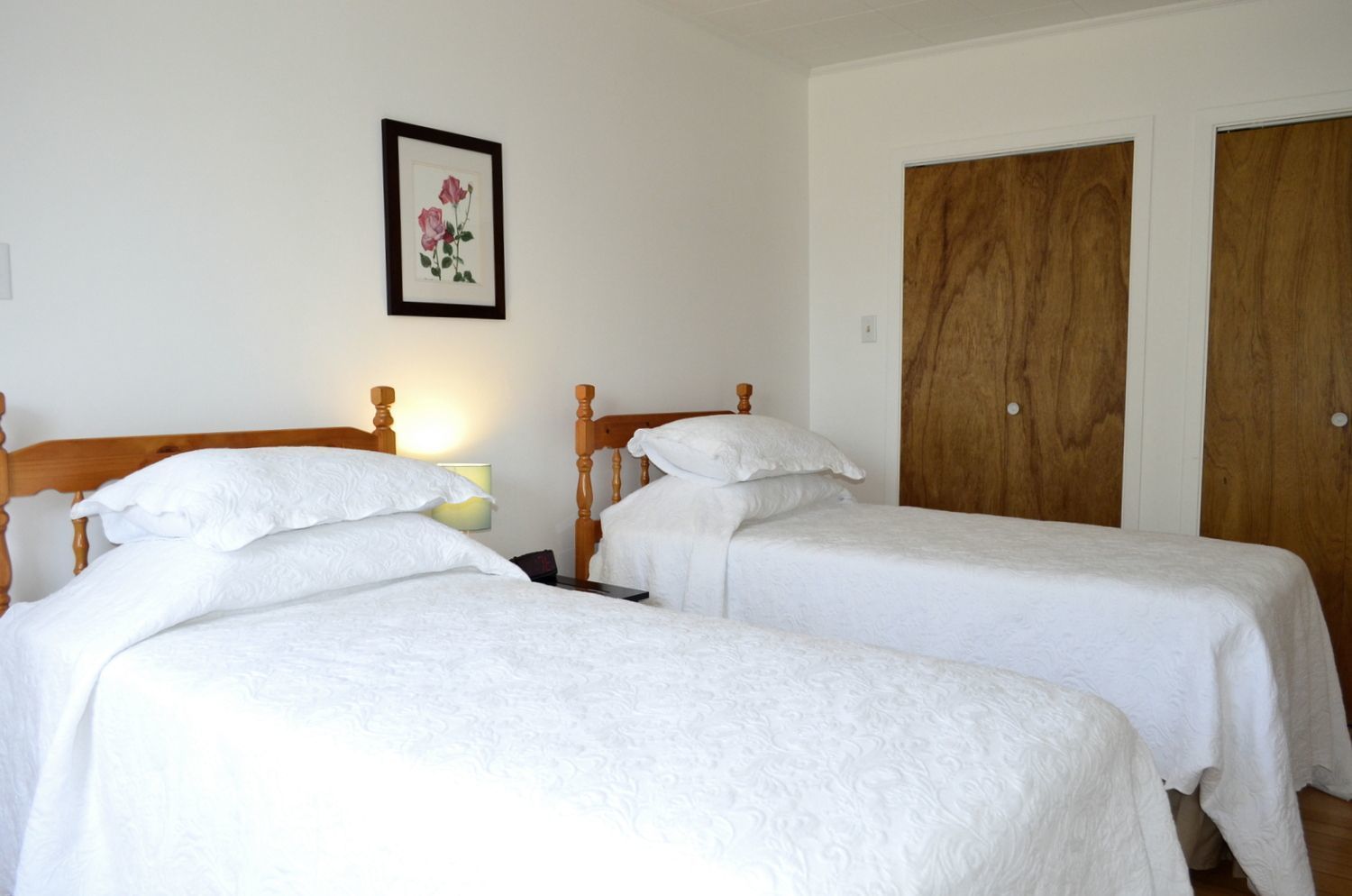 A room with two single beds, wooden headboards, white bedding, a framed floral artwork on the wall, and wooden doors.