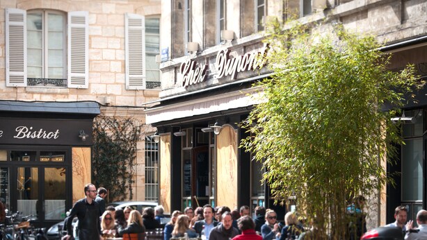 An outdoor café with patrons seated at tables, a waiter walking by, and a building with a sign that reads "Café Dupont". - Une Chambre chez Dupont (Bordeaux)