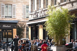 Un café al aire libre con clientes sentados en mesas, un camarero pasando y un edificio con un cartel que dice "Café Dupont".