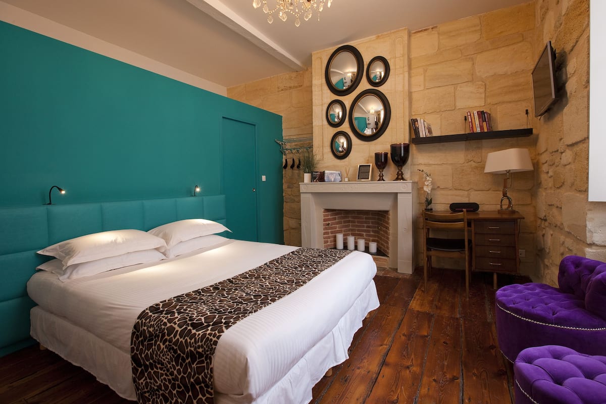 A bedroom with a large bed, a teal accent wall, a fireplace, a desk, and a purple armchair.