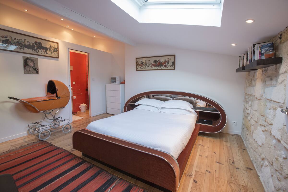 A modern bedroom with a skylight, a wooden bed, a red door, and a wall-mounted shelf with books.