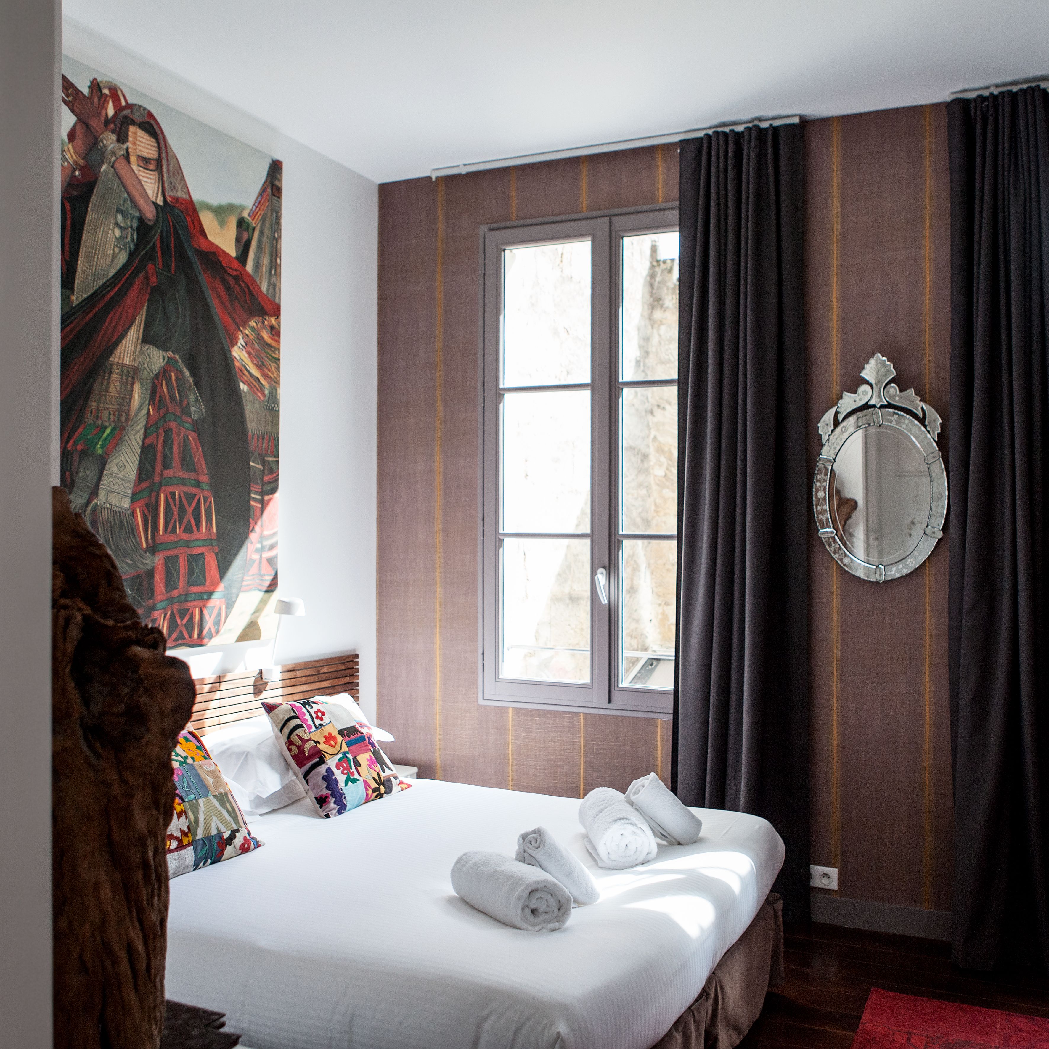 A hotel room with a large bed, a window with curtains, a mirror, and a colorful tapestry on the wall.