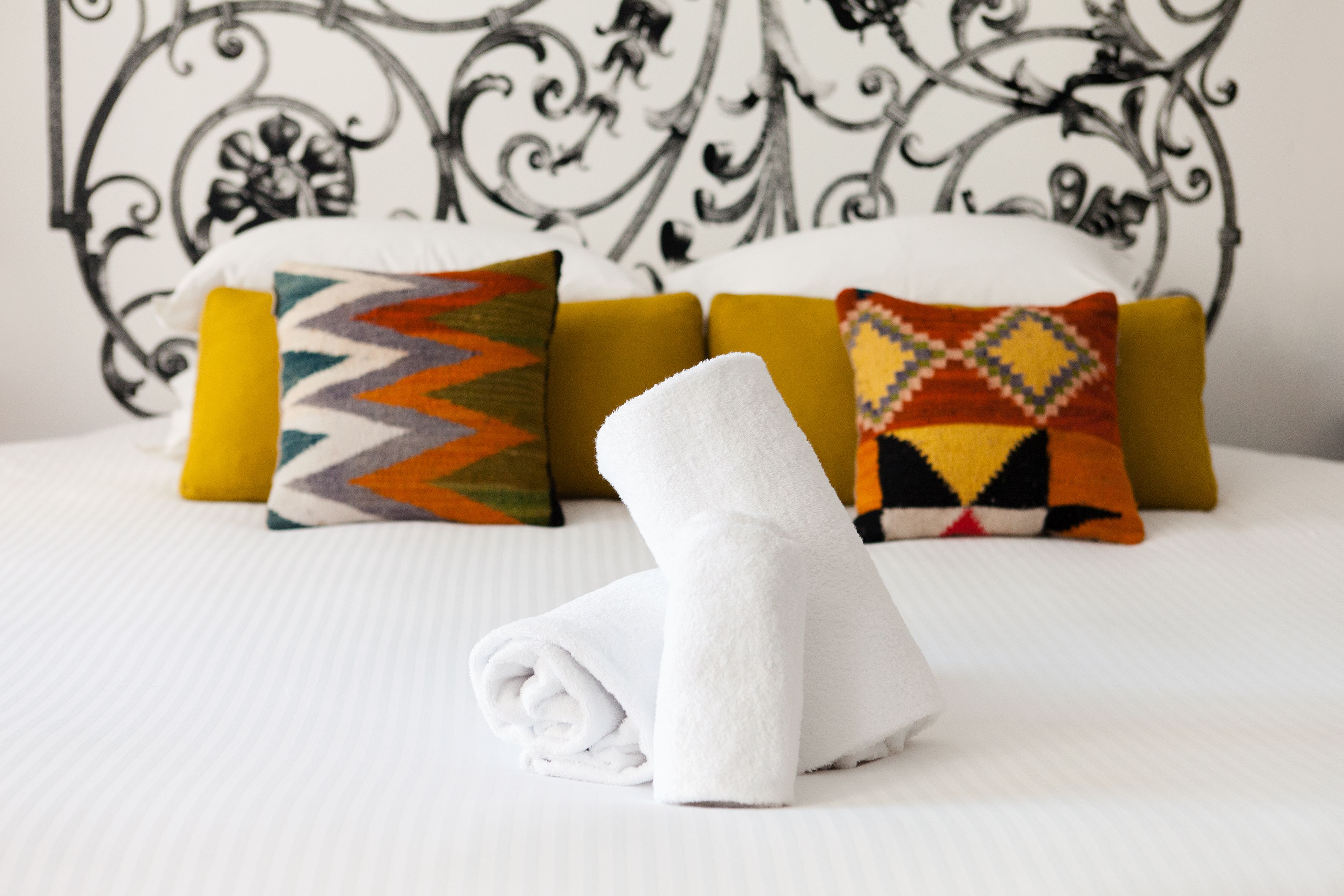 Un lit bien fait, avec une serviette blanche roulée, des coussins colorés et une tête de lit décorative en métal.