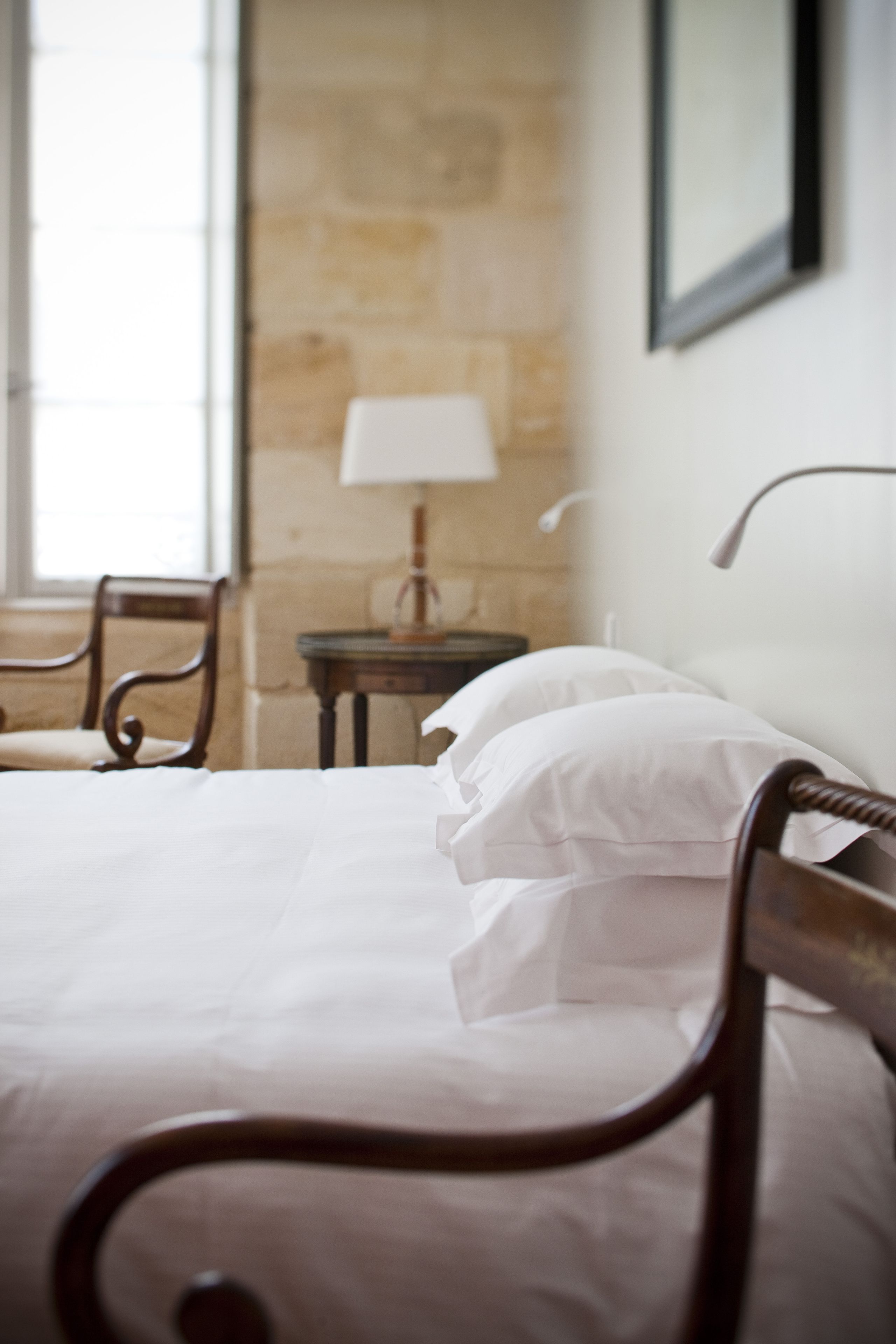 A neatly made bed with white linens and a wooden headboard, a small bedside table with a lamp, and a chair in a room with stone walls.
