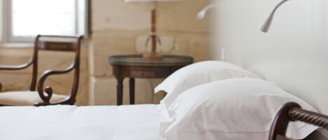A neatly made bed with white linens and a wooden headboard, a small bedside table with a lamp, and a chair in a room with stone walls.