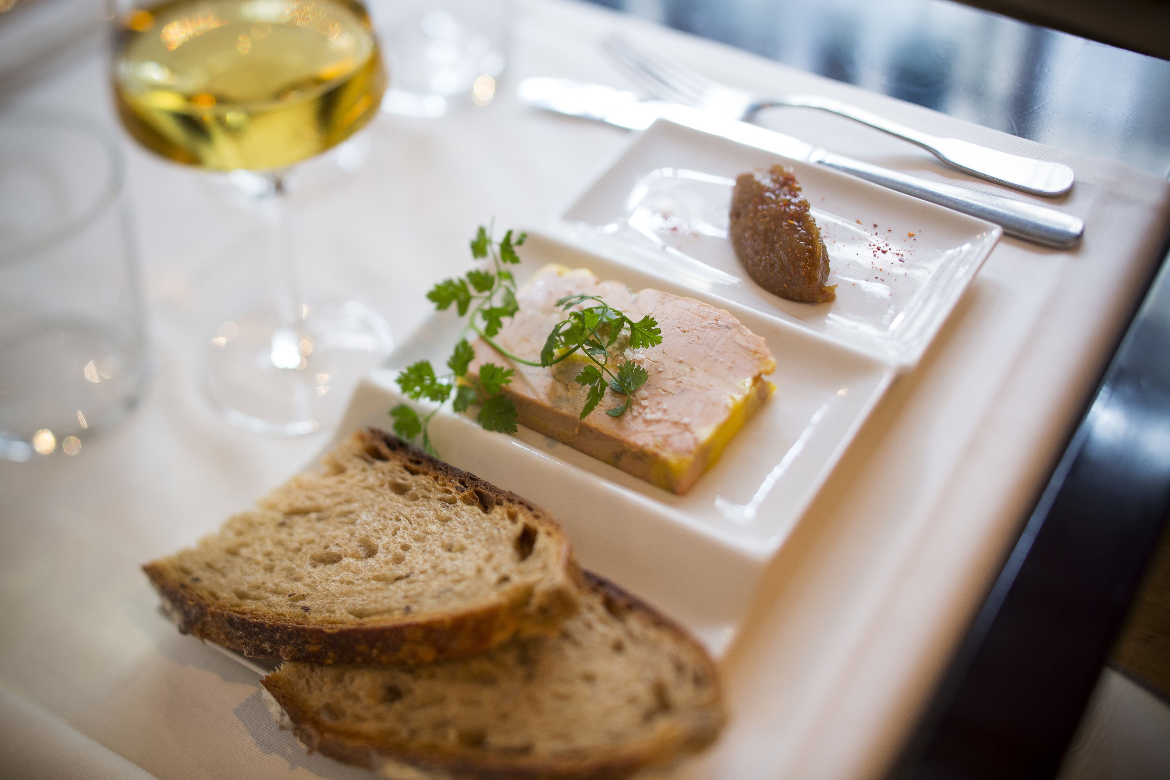 Een eettafel met een wit bord met een sneetje brood, een stuk vlees en een glas witte wijn.