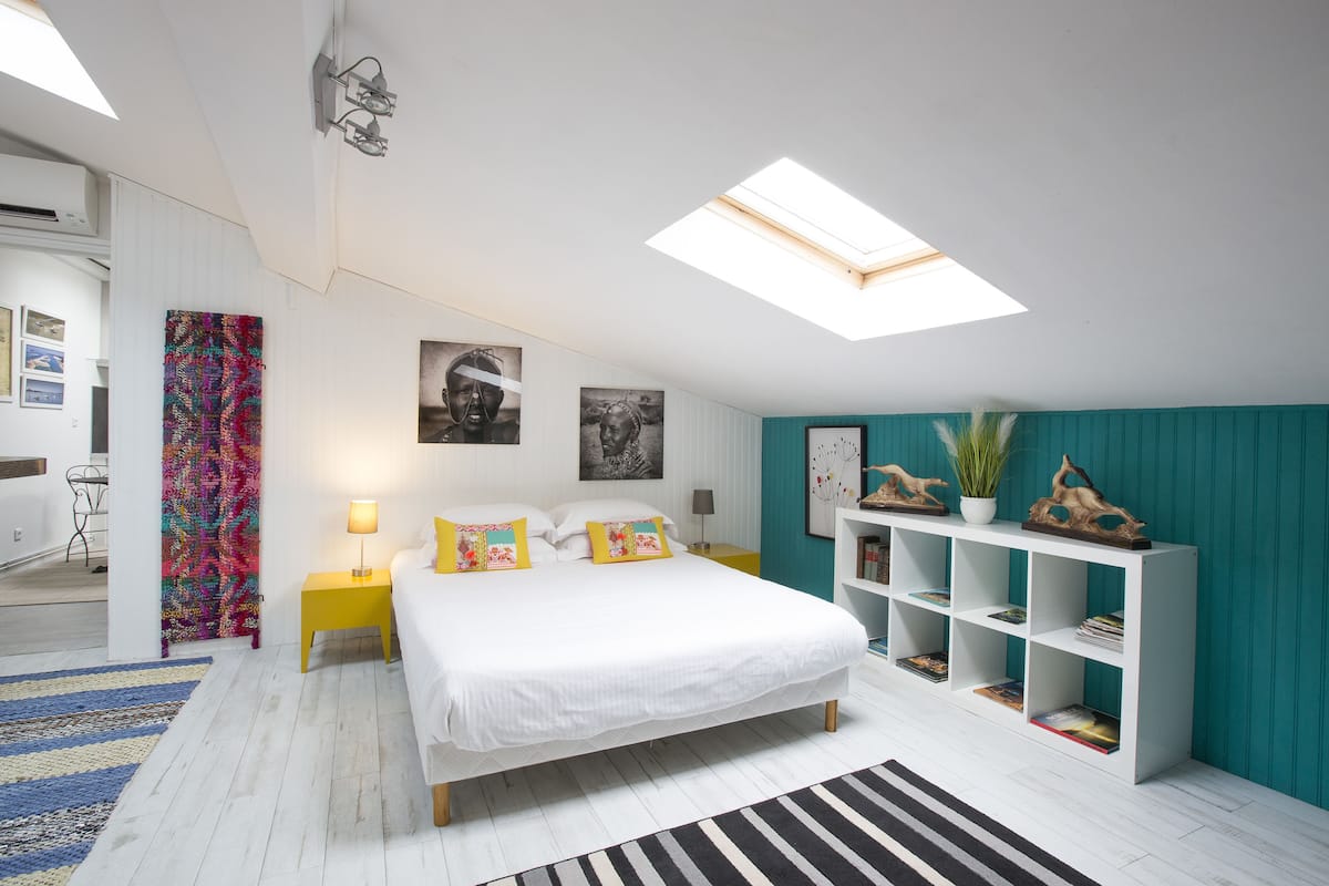 A modern bedroom with a skylight, a bed with white linens, a yellow nightstand, a teal accent wall, and a white bookshelf.