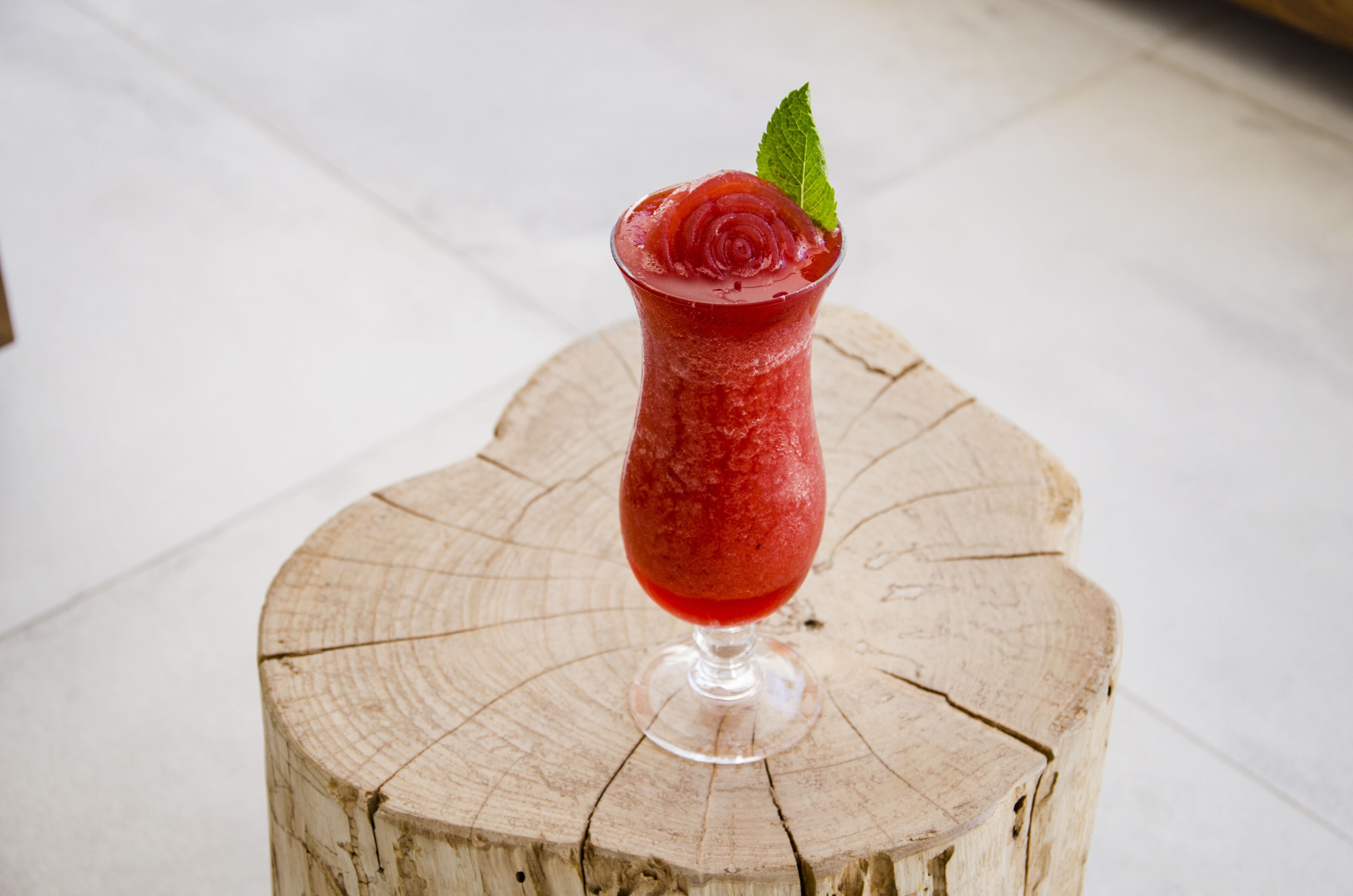 Un cóctel rojo en un vaso con diseño de rosa, adornado con una hoja de menta, sobre un tocón de madera.