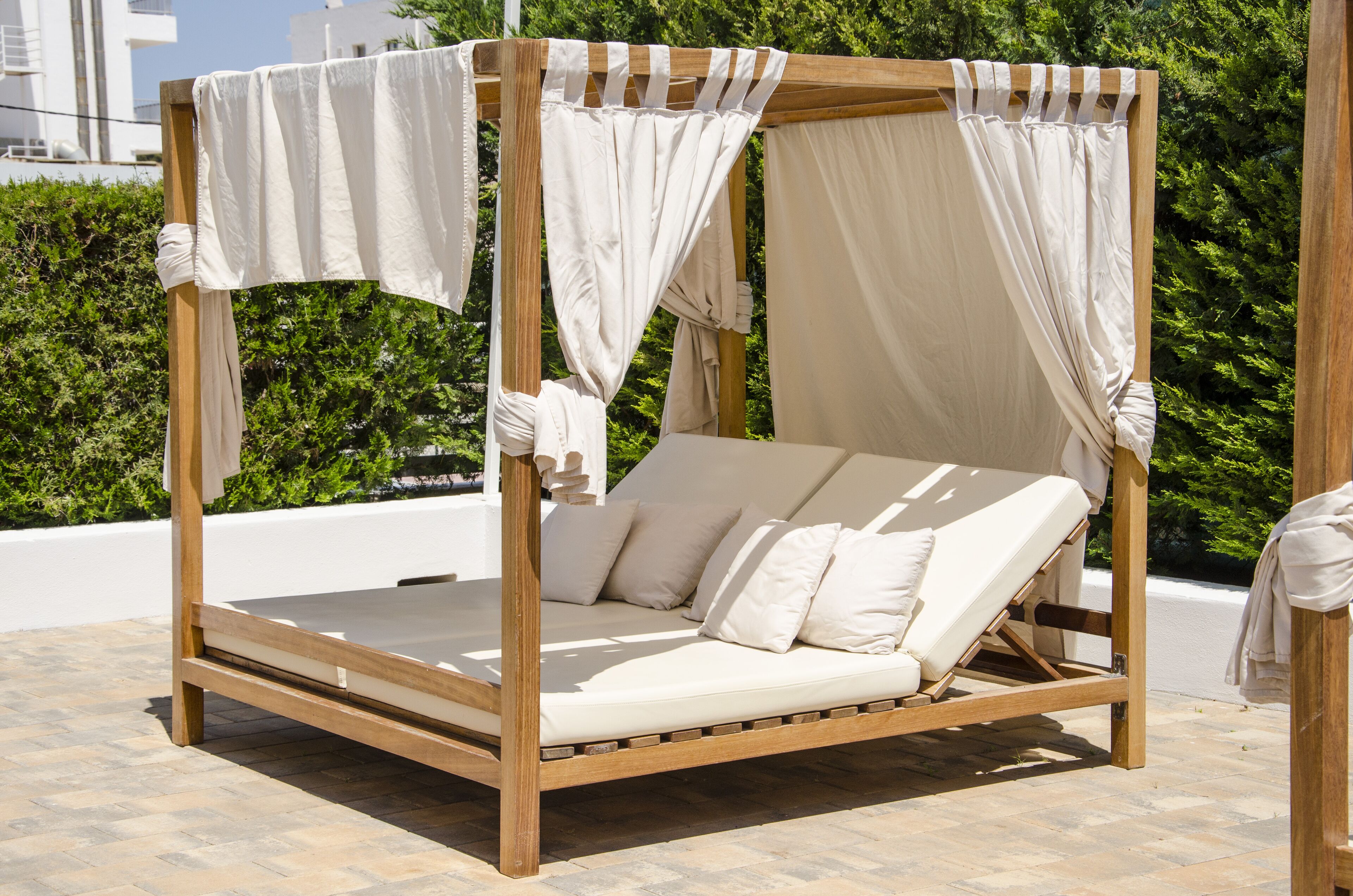 Un lit à baldaquin en bois, surmonté d’un dais, avec du linge de lit blanc, placé sur une terrasse carrelée d’où l’on peut admirer un paysage verdoyant et des bâtiments en arrière-plan.
