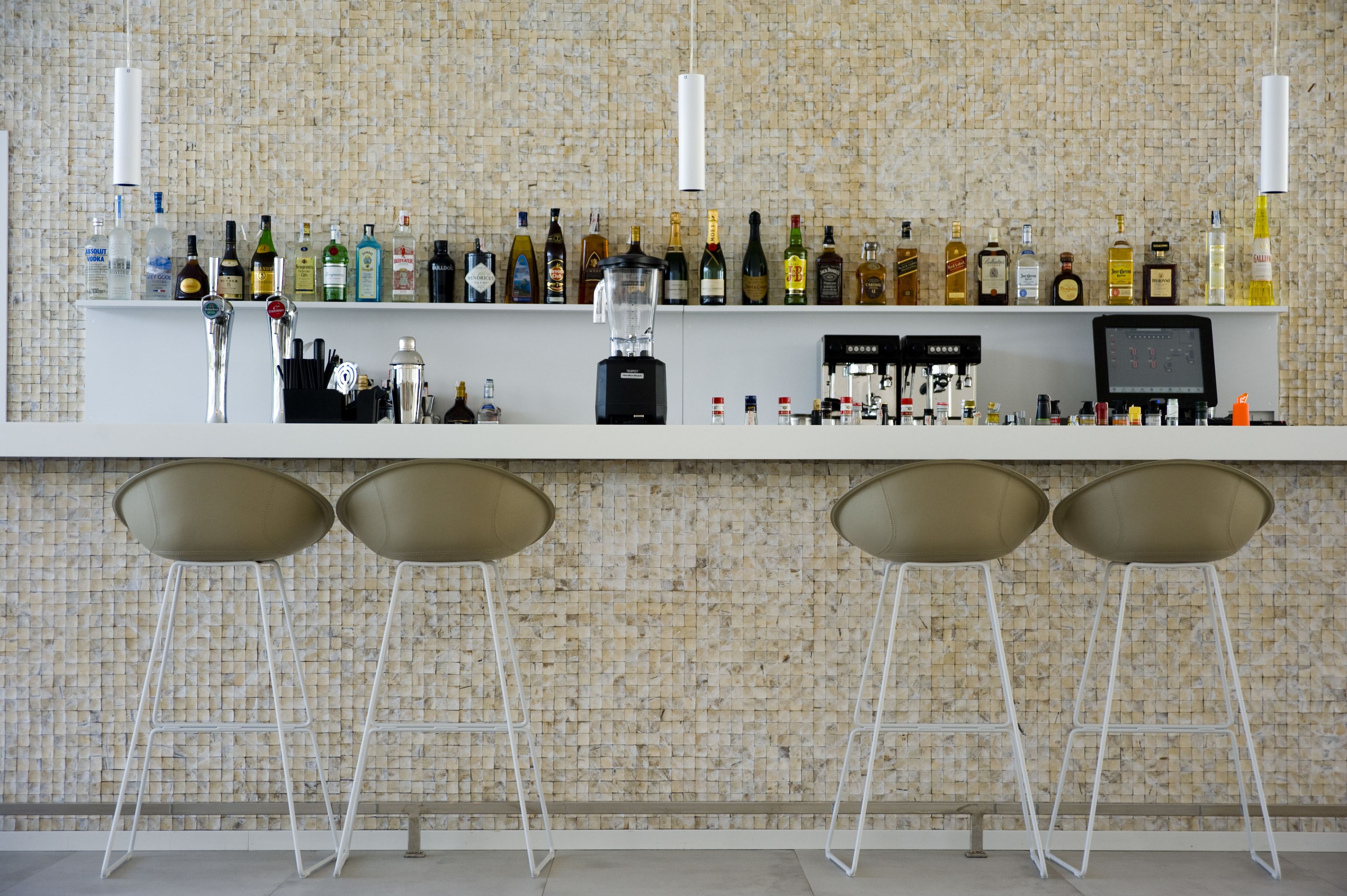 Zona de bar con diversas botellas de licor, una licuadora y una pantalla digital. Hay tres taburetes frente a la barra.