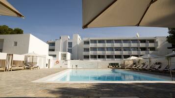 Una piscina con sillones de descanso y sombrillas, rodeada por un edificio moderno de varios pisos y balcones.