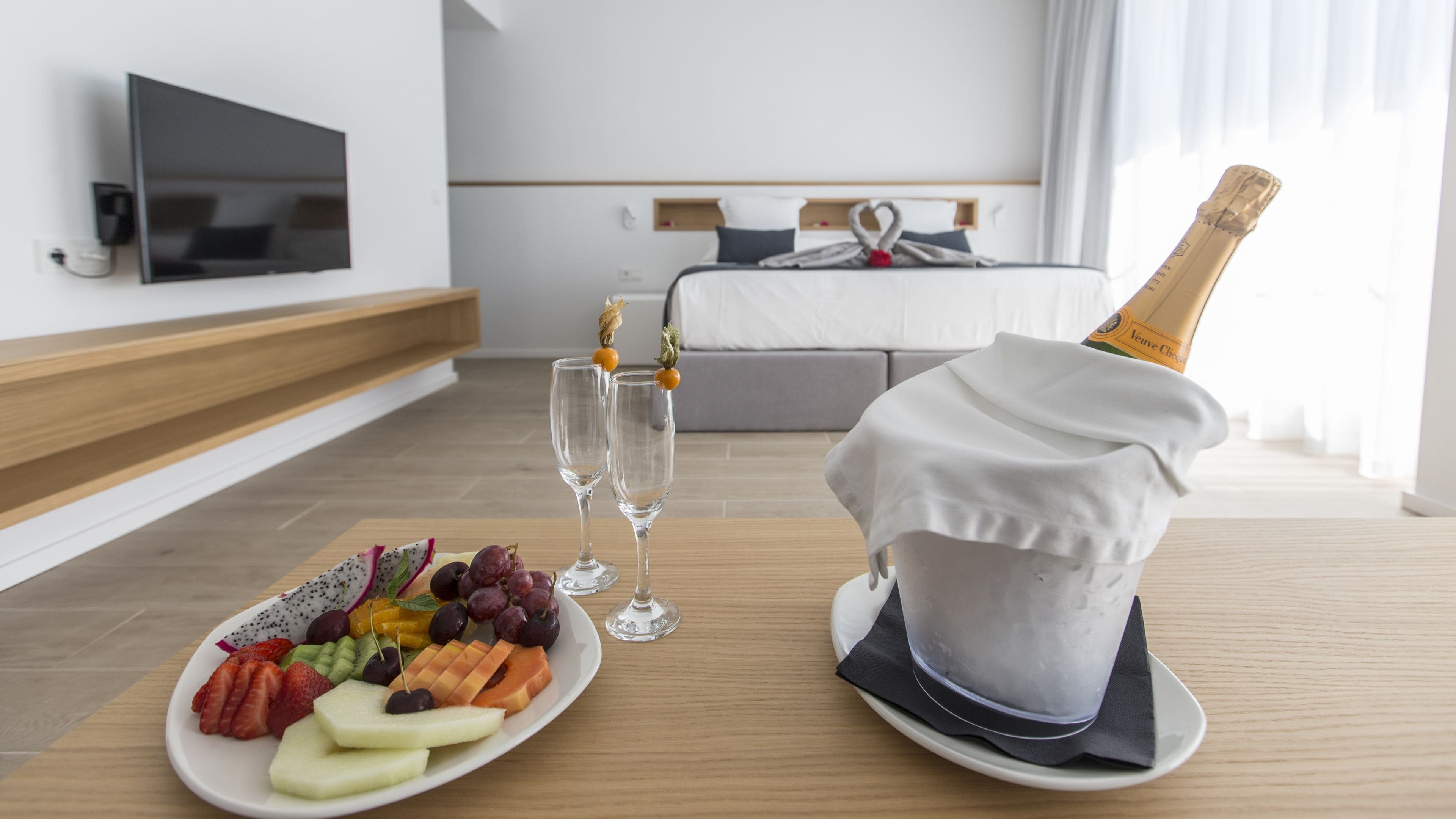 A hotel room with a bed, a TV, a fruit platter, two glasses, and a bottle of champagne.