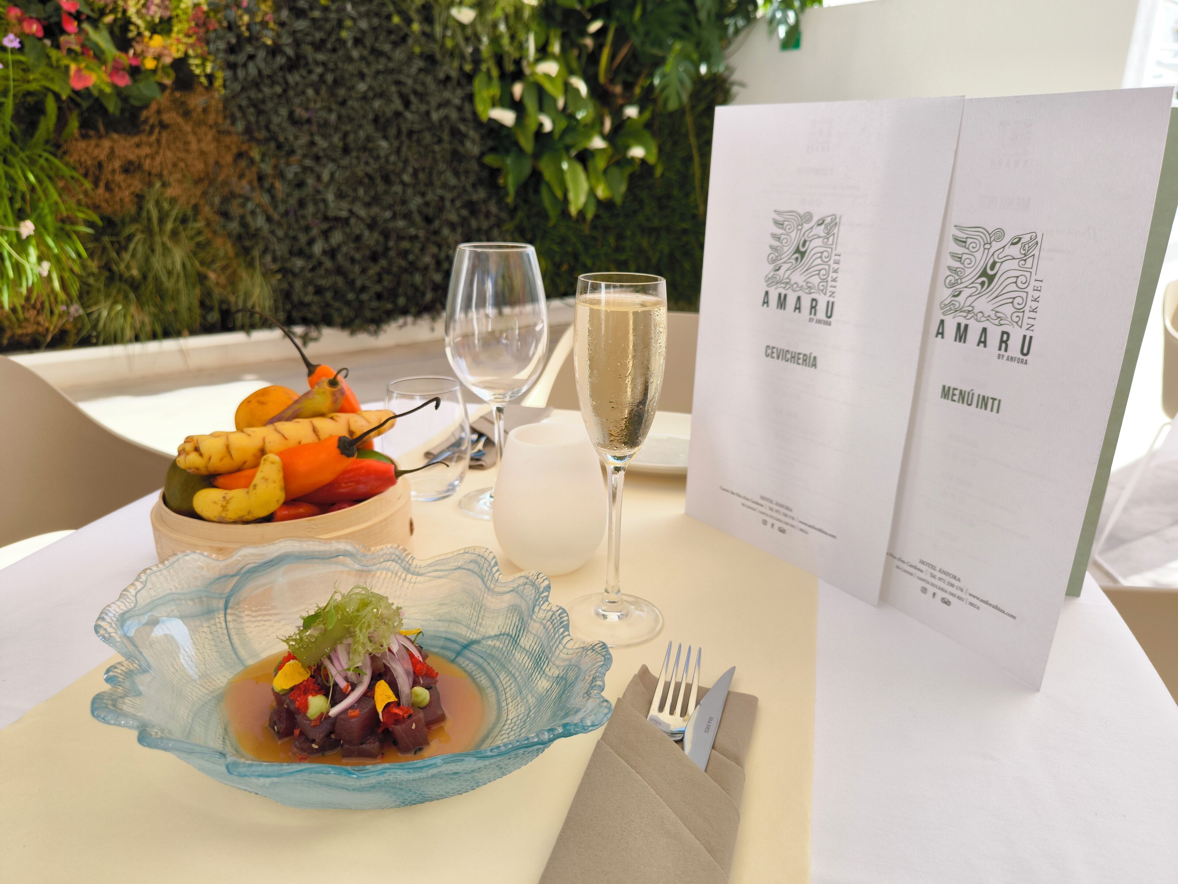 Una mesa servida con una copa de champán, un cuenco de fruta, un plato de ceviche y un menú.