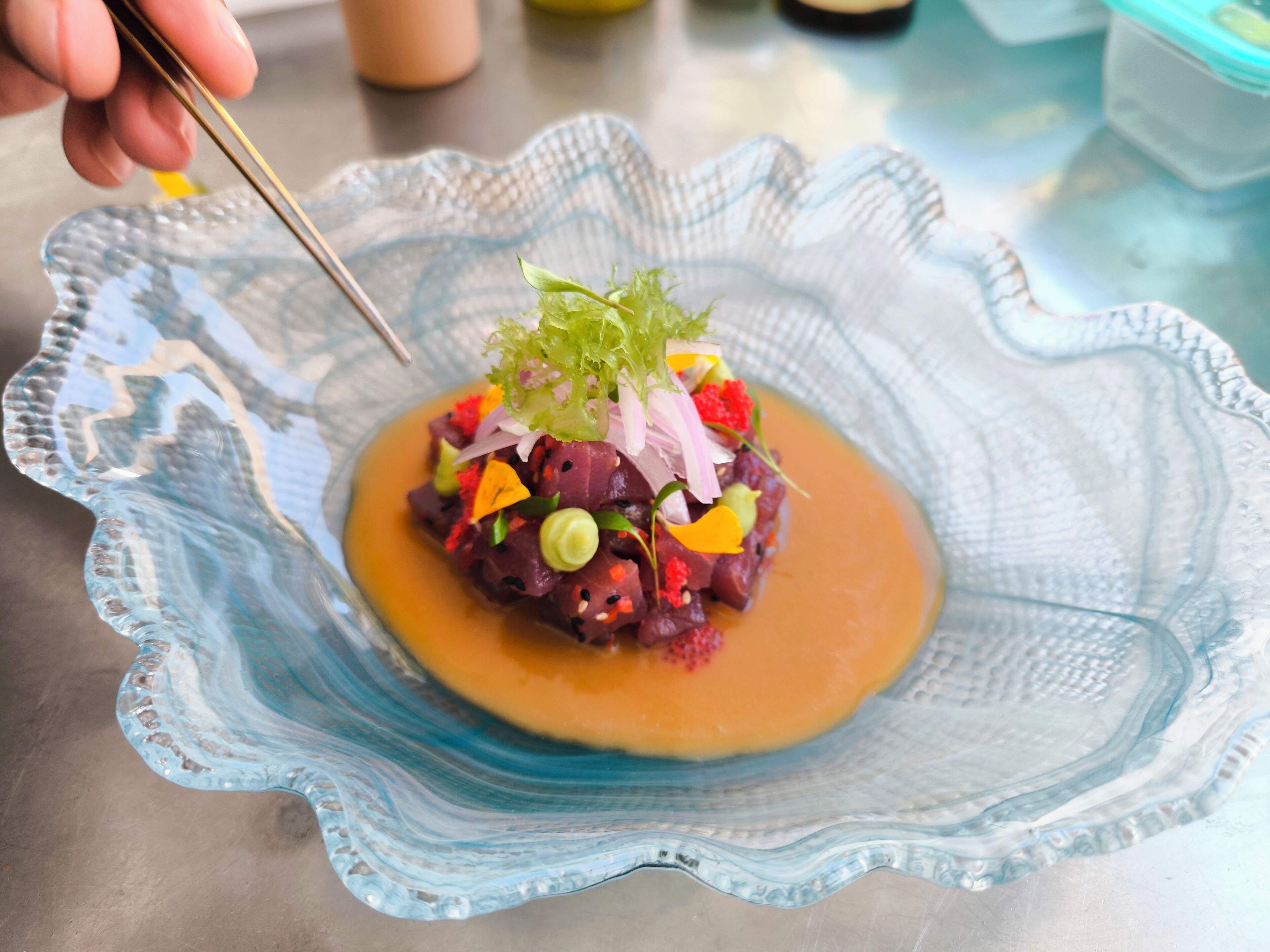 A glass bowl with a decorative edge containing a dish garnished with green leaves, red and yellow edible flowers, and a sauce.