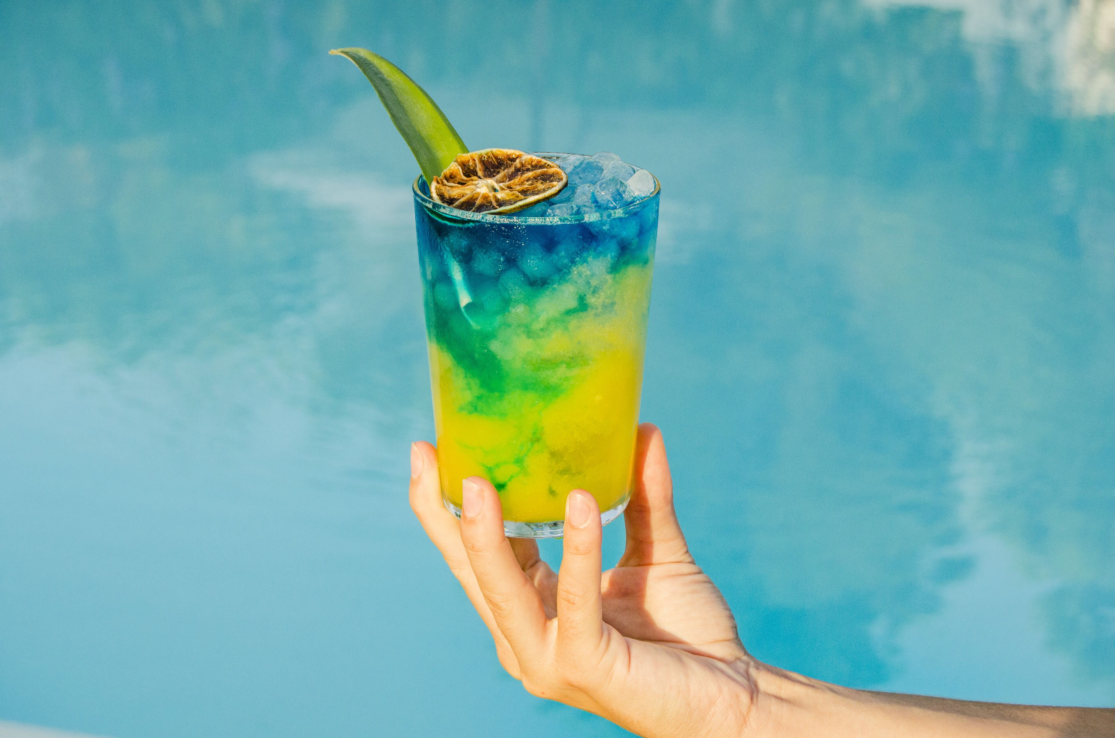 Una mano sostiene un vaso con una bebida tropical, hielo y una rodaja de fruta, sobre un fondo azul.