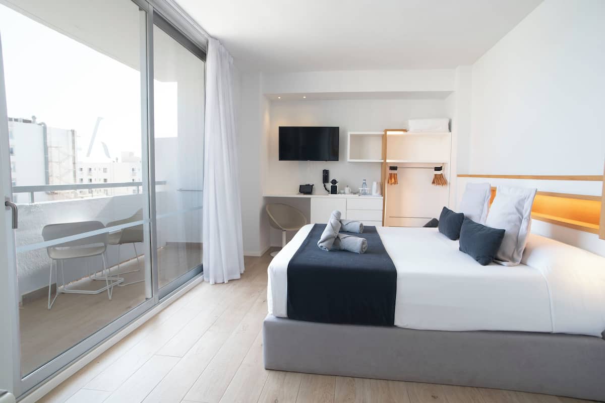 A modern hotel room with a large bed, a desk, a chair, a TV, and a balcony with a city view.