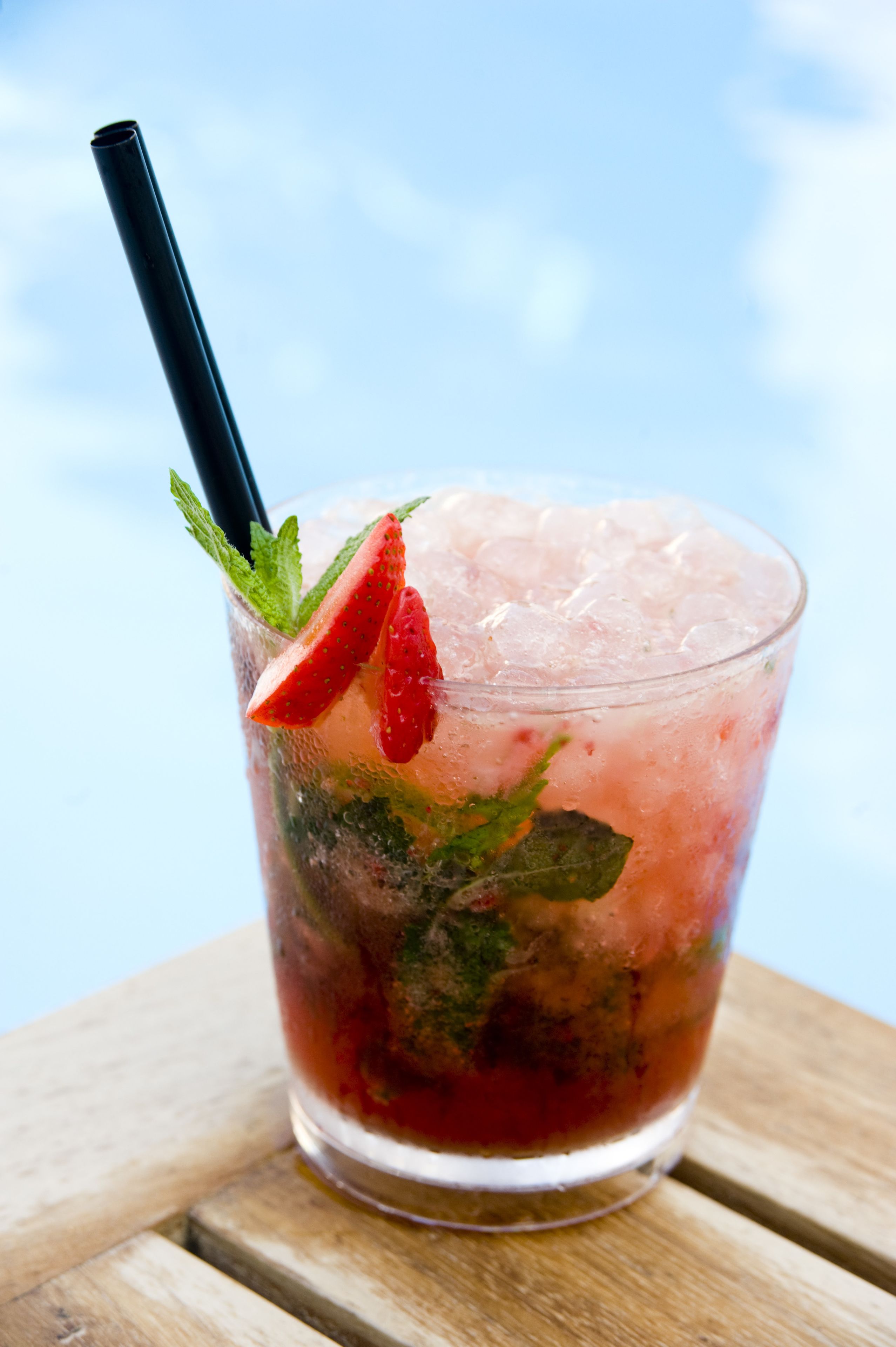 A glass of iced beverage with a straw, garnished with a strawberry slice and mint leaves, placed on a wooden surface.