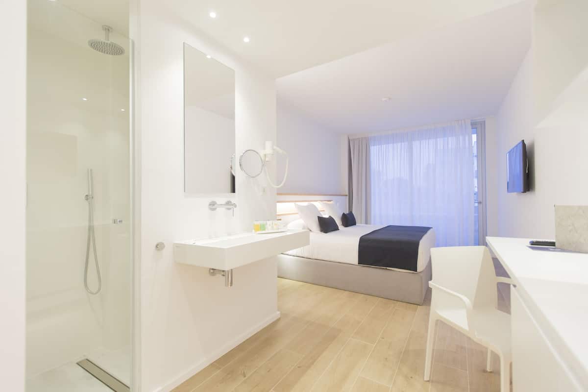 A modern hotel room with a glass-enclosed shower, a white sink, a bed with a black throw, and a flat-screen TV mounted on the wall.